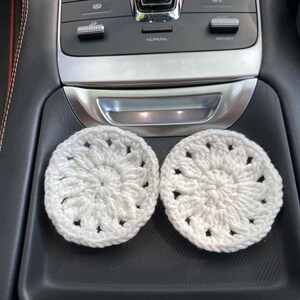 May include: Two white crocheted coasters with a floral design. The coasters are sitting in the cup holders of a car.