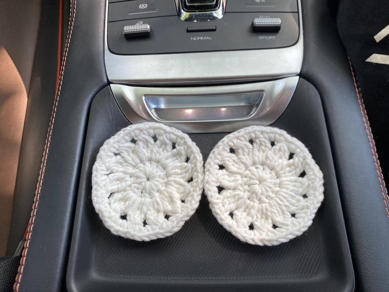 May include: Two white crocheted coasters with a floral design. The coasters are sitting in the cup holders of a car.