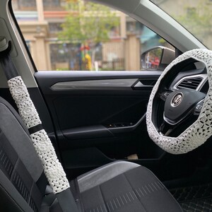 May include: White crocheted steering wheel cover and seatbelt covers in a car interior. The steering wheel cover has a crocheted design with a white background and a black Volkswagen logo in the center.