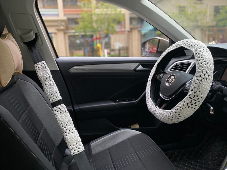 May include: White crocheted steering wheel cover and seatbelt covers in a car interior. The steering wheel cover has a crocheted design with a white background and a black Volkswagen logo in the center.
