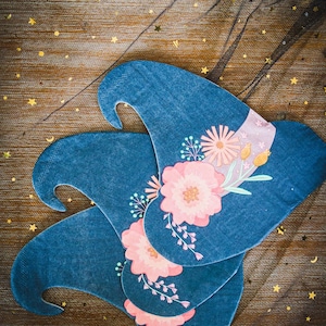 May include: Three blue witch hats with floral designs. Each hat features a pink and white flower arrangement with green leaves and a lavender band. The hats are arranged on a gold and black star-patterned surface.