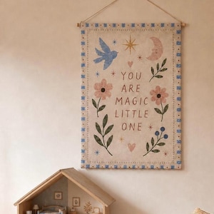 May include: A rectangular fabric wall hanging featuring the text YOU ARE MAGIC LITTLE ONE in brown letters. The design includes a blue bird, a pink crescent moon, gold stars, pink flowers, green leaves, and blue berries, all framed by a blue and white border.