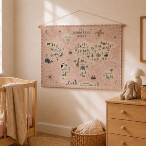 May include: A rectangular wall hanging featuring a world map on a dusty pink fabric background. The map displays the text Our Wonderful World, along with various illustrations of animals, landmarks, and trees across the continents, suspended by a wooden rod and a thin string.