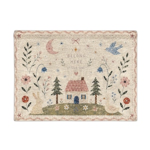 May include: A rectangular woven tapestry featuring a cream background with a central house, pine trees, a rabbit, and a duck. The text reads YOU BELONG HERE LITTLE ONE. The design includes a pink moon, blue bird, flowers, and a scalloped border.