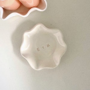 May include: A small, cream-coloured ceramic dish with a wavy, flower-like shape. The dish has the initials "E + M" stamped in the centre. Another white dish with pink objects is visible in the background.