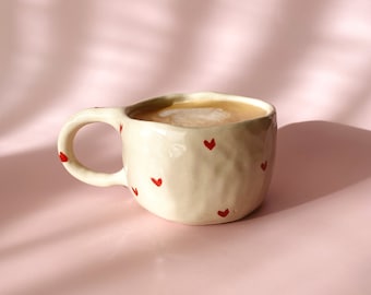 May include: A cream-colored ceramic mug filled with a light brown beverage. The mug has a textured surface and is decorated with small red hearts. The mug is sitting on a pink surface.