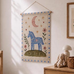 Puede incluir: Un tapiz rectangular de tela con la ilustración de un caballo azul sobre una colina verde. El diseño incluye una luna creciente rosa, estrellas amarillas y flores rosas. El borde tiene un patrón de cuadros azules y blancos.