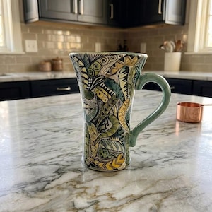 May include: A tall ceramic mug featuring a complex, hand-drawn pattern of interlocking shapes and lines in shades of olive green, mustard yellow, and black. The mug has a smooth, light green handle and a flared rim, showcasing intricate details.