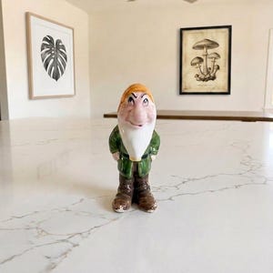 May include: A ceramic figurine of a dwarf with a large nose, a long white beard, an orange hat, a dark green jacket, and brown boots. The figure has a glossy finish and stands in an upright position with hands placed behind the back.