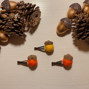 Puede incluir: Tres pinzas para el pelo de fieltro en forma de bellota, una amarilla, dos naranjas, con tapas de fieltro marrón. Las pinzas están sobre una superficie blanca con piñas doradas.