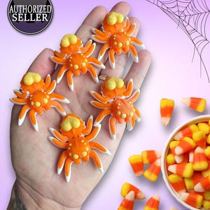 Puede incluir: Cinco arañas impresas en 3D de color naranja y blanco con ojos amarillos. Las arañas están sentadas en una mano. También hay piezas de caramelo de maíz naranja y amarillo en el fondo.