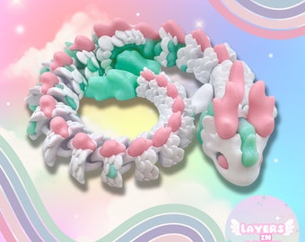 Può includere: Una statuetta di drago a strati di colore bianco, rosa e verde. Il drago è arrotolato in un cerchio con la testa appoggiata sulla coda. La statuetta è realizzata in un materiale morbido e flessibile e ha un aspetto fantasioso e giocoso.