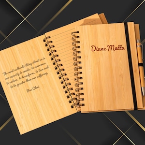 May include: A wooden spiral-bound notebook with a pen. The cover is engraved with the name "Diane Malla". The open pages display a quote by Ben Ohri. The notebook is made of light-colored wood.