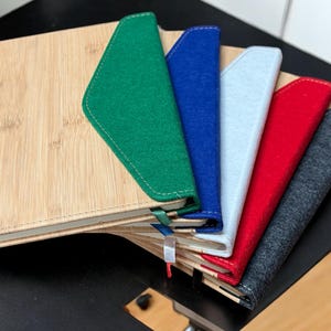 Puede incluir: Una pila de seis cuadernos con cubiertas de fieltro de diferentes colores. El cuaderno de arriba tiene una cubierta de bambú natural. Las cubiertas son verdes, azules, blancas, rojas, grises y negras.