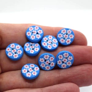 May include: A collection of seven round, bright blue buttons, each featuring a repeating pattern of small, five-petalled white flowers with pink centres. Each button has two central holes for attachment, arranged in a circular, flat design.