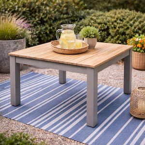 May include: A square outdoor coffee table with a light wood top and gray legs. A woven tray holds a pitcher of lemonade and two glasses. A small potted plant sits on the table. The table rests on a blue and white striped rug.