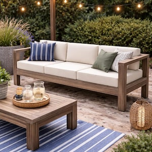 Puede incluir: Conjunto de muebles de patio exterior de madera con un sofá con cojines color crema y cojines decorativos. También hay una mesa de centro a juego y una linterna tejida. La escena está ambientada en una terraza de madera con una alfombra a rayas azules y blancas.
