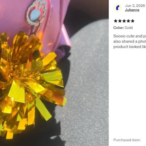 May include: A metallic gold cheerleader pom-pom keychain attached to a pink bag. The pom-pom is constructed from numerous thin, reflective strips of gold material that fan out in a spherical shape, secured by a silver-toned metal ring attachment.