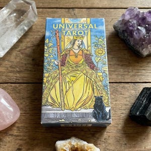May include: A Universal Tarot deck box featuring a queen in a yellow gown, a black cat, and the text "UNIVERSAL TAROT." The box is surrounded by crystals: clear quartz, amethyst, rose quartz, citrine, and black tourmaline. The items are arranged on a wooden surface.
