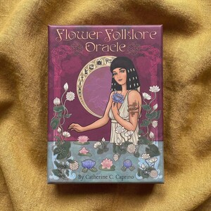 May include: A rectangular box for the "Flower Folklore Oracle" deck. The box features an illustration of a woman holding a blue flower, surrounded by flowers and water lilies. The text "Flower Folklore Oracle" and the author's name are visible.