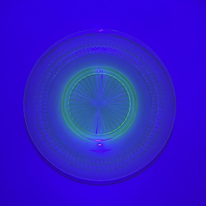 May include: A clear glass plate with a central, radiating design and a patterned rim. The plate glows with a neon green hue in the center, surrounded by a blue glow, set against a deep blue background.