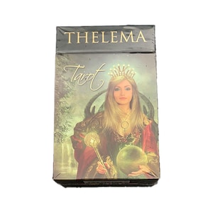 May include: A tarot deck box with a gold and black design. The box features a woman with long blonde hair wearing a crown and a robe. The text "THELEMA Tarot" is printed on the box.