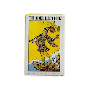 May include: A pocket edition of The Rider Tarot Deck. The box features a yellow background with an illustration of a figure with a staff, a dog, and a sun. The figure is wearing a patterned outfit and boots. The text "THE RIDER TAROT DECK" is at the top.