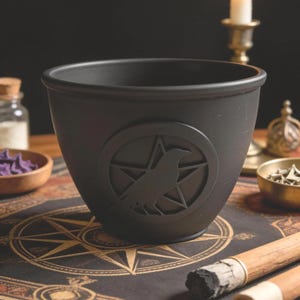 May include: A black cauldron with a raven and pentagram design. The cauldron is round with a wide rim and a slightly flared base. The raven is centered within a five-pointed star. The cauldron is sitting on a table with other items.