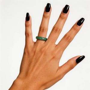 May include: A green jade ring is worn on a hand with black nail polish. The ring is a solid band of jade, showcasing a natural, mottled green color. The hand is positioned against a white background, highlighting the ring's color and design.