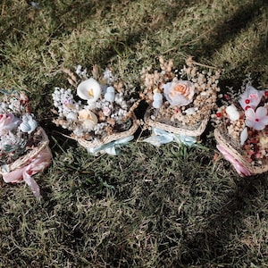 May include: Four small baskets filled with dried flowers and decorative elements. The baskets are arranged in a row on a bed of green grass. Each basket has a different color ribbon tied around it.