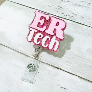 Op de afbeelding: Een roze en glinsterende ER Tech badge reel met witte letters. De badge reel heeft een doorzichtige plastic band en een metalen clip om aan kleding te bevestigen. De achtergrond is een lichte houtnerf.