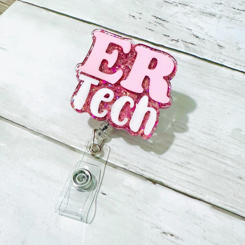 Op de afbeelding: Een roze en glinsterende ER Tech badge reel met witte letters. De badge reel heeft een doorzichtige plastic band en een metalen clip om aan kleding te bevestigen. De achtergrond is een lichte houtnerf.