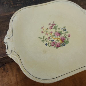 May include: Cream-colored serving tray with a floral design. The tray features a central arrangement of pink and blue flowers with green leaves and a gold-colored branch. The tray has a scalloped edge with a black trim and decorative detailing.