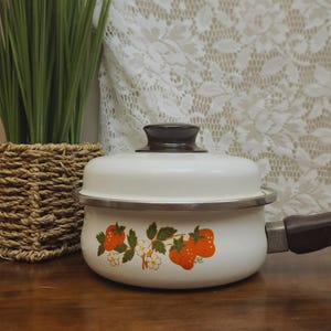 May include: A white enameled metal saucepan with a matching lid, featuring a brown handle and knob. The exterior is decorated with a graphic print of red strawberries, green leaves, and white blossoms. The pot has a silver-toned metal rim.