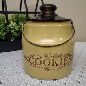 May include: A cylindrical ceramic cookie jar with a pale yellow glaze and a dark brown lid. The front features the word COOKIES in brown lettering, surrounded by a decorative brown floral and leaf pattern. A thin metal wire handle is attached.