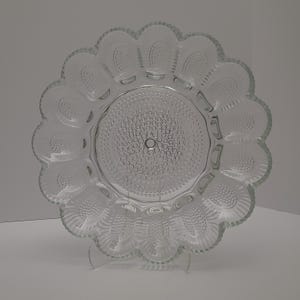 May include: Clear glass serving dish with a scalloped edge and a central circular design. The dish has a textured surface with a pattern of small circles and is supported by a clear stand. Ideal for serving appetizers or desserts.
