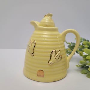 May include: A pale yellow ceramic creamer shaped like a beehive, featuring a ribbed texture, a small handle, and a pointed lid. The exterior is decorated with two gold-painted flying birds and a small gold-painted arched doorway at the base.