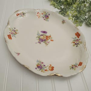 May include: An oval, off-white serving platter with a scalloped edge and gold trim. The platter is decorated with clusters of colorful flowers in shades of orange, purple, yellow, and blue. A vintage piece.