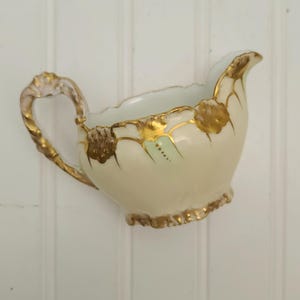 May include: Cream-colored porcelain creamer with gold trim and accents. The handle is twisted and gold-toned. The rim and base have a scalloped design. The creamer has a small spout for pouring. The creamer has a light green interior.