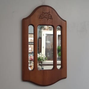 May include: A wall-mounted mirror featuring a medium-toned wooden frame with an arched top and a carved floral motif. The frame contains three vertical glass panels, with a larger central mirror flanked by two narrower, elongated side mirrors.