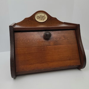 May include: A brown wooden bread box with a slanted front door and a dark circular knob. The top features an arched back panel with an oval cream-colored plaque centered at the top, displaying the word Bread in dark brown script lettering.