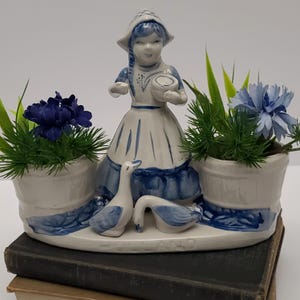 May include: A ceramic planter featuring a Dutch girl figurine in a blue and white dress and bonnet, holding a bowl. Two geese are at the base. Two white planters with blue trim hold blue flowers and greenery. The planter sits on a stack of books.