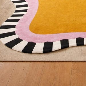 May include: A colorful rug with an abstract design. The rug features a mustard yellow center, a pink border, and a black and white striped edge. The rug is placed on a neutral-colored rug and hardwood floor.