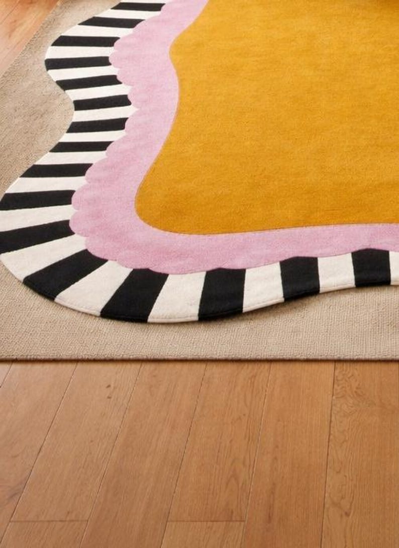 May include: A colorful rug with an abstract design. The rug features a mustard yellow center, a pink border, and a black and white striped edge. The rug is placed on a neutral-colored rug and hardwood floor.