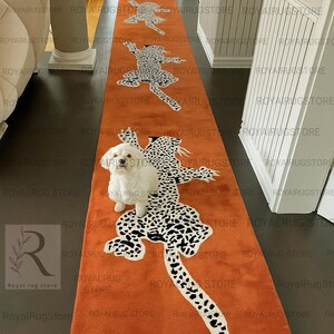 Puede incluir: Una alfombra de pasillo naranja con un patrón repetido de figuras de leopardo estilizadas en blanco y negro. La alfombra rectangular parece estar hecha de un material suave, adecuada para un pasillo o entrada.