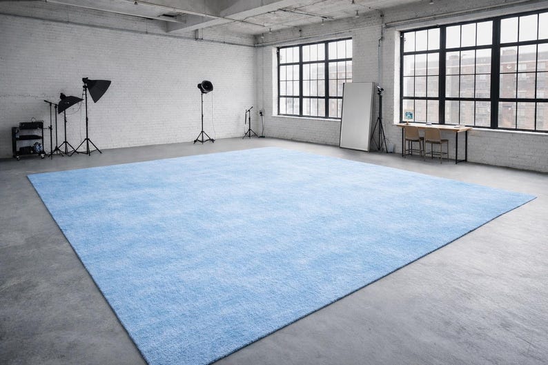 May include: A large, rectangular, light blue rug dominates a concrete floor in a studio setting. The rug's texture is visible, and the room features large windows, lighting equipment, and a white brick wall.