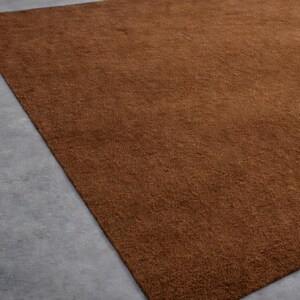 May include: A large, rectangular brown rug with a textured surface. The rug is placed on a gray concrete floor, with a portion of the rug filling the frame. The rug's color is a warm, earthy tone.