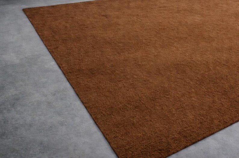 May include: A large, rectangular brown rug with a textured surface. The rug is placed on a gray concrete floor, with a portion of the rug filling the frame. The rug's color is a warm, earthy tone.