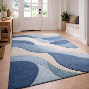 Può includere: Un tappeto rettangolare con un design a onde in tonalità di blu e beige. Il tappeto è posizionato su un pavimento in legno in un ingresso luminoso. Il design presenta linee curve e fluide, creando un senso di movimento.