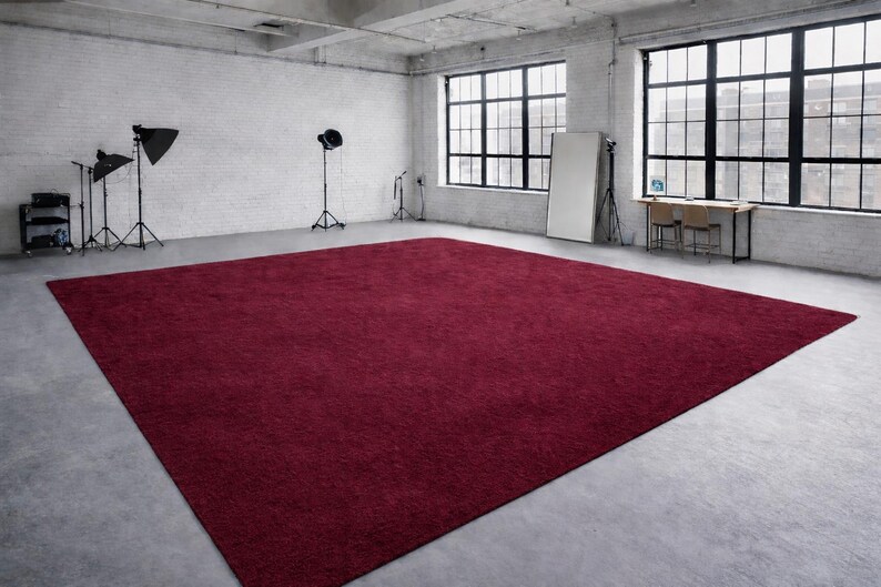 May include: A large, rectangular, burgundy-colored rug dominates a studio space. The rug is placed on a concrete floor, and the room features large windows, studio lights, and a white brick wall.